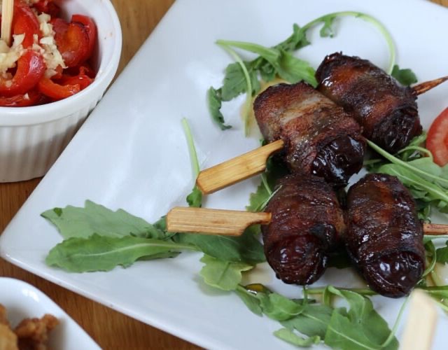 Drei in Speck gewickelte Datteln am Holzspieß auf Rucola, dunkel gebraten.