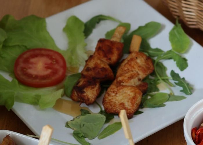 Zwei gegrillte Hähnchenspieße auf einem weißen Teller mit Rucola, Blattsalat und einer Tomatenscheibe.