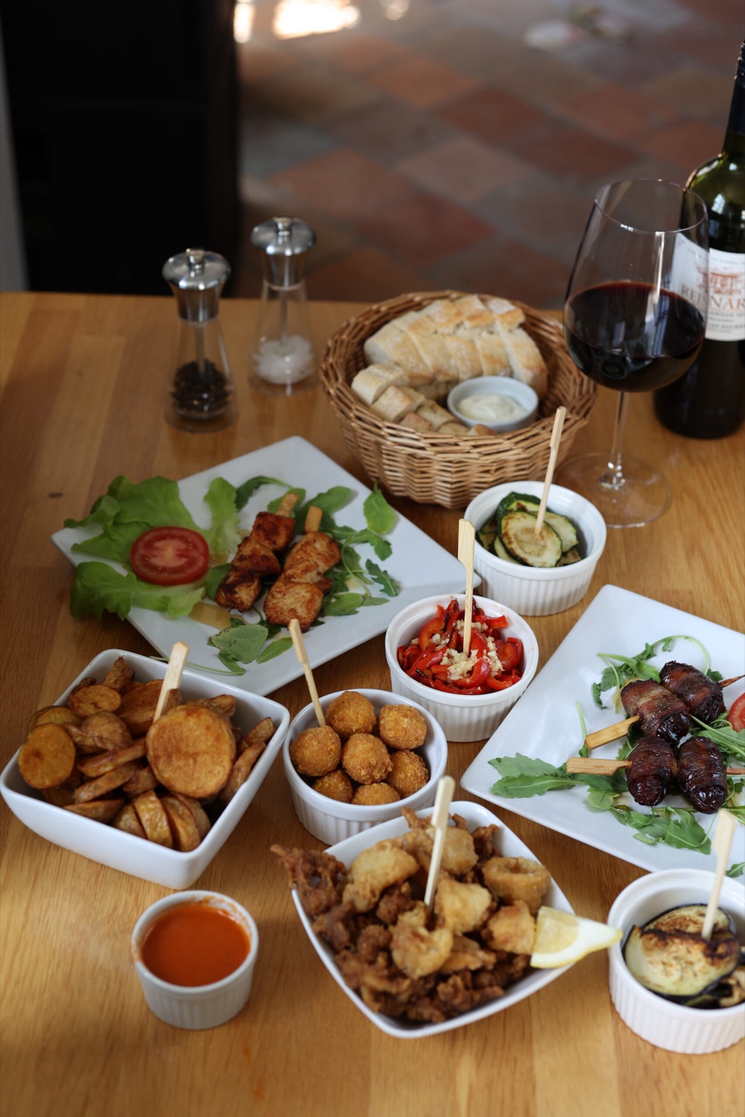 Ein gedeckter Holztisch im El Moro mit Hähnchenspießen, Datteln im Speckmantel, frittierten Calamari, Kroketten, Bratkartoffeln, gegrillter Paprika und Zucchini, dazu Brotkorb und ein Glas Rotwein.